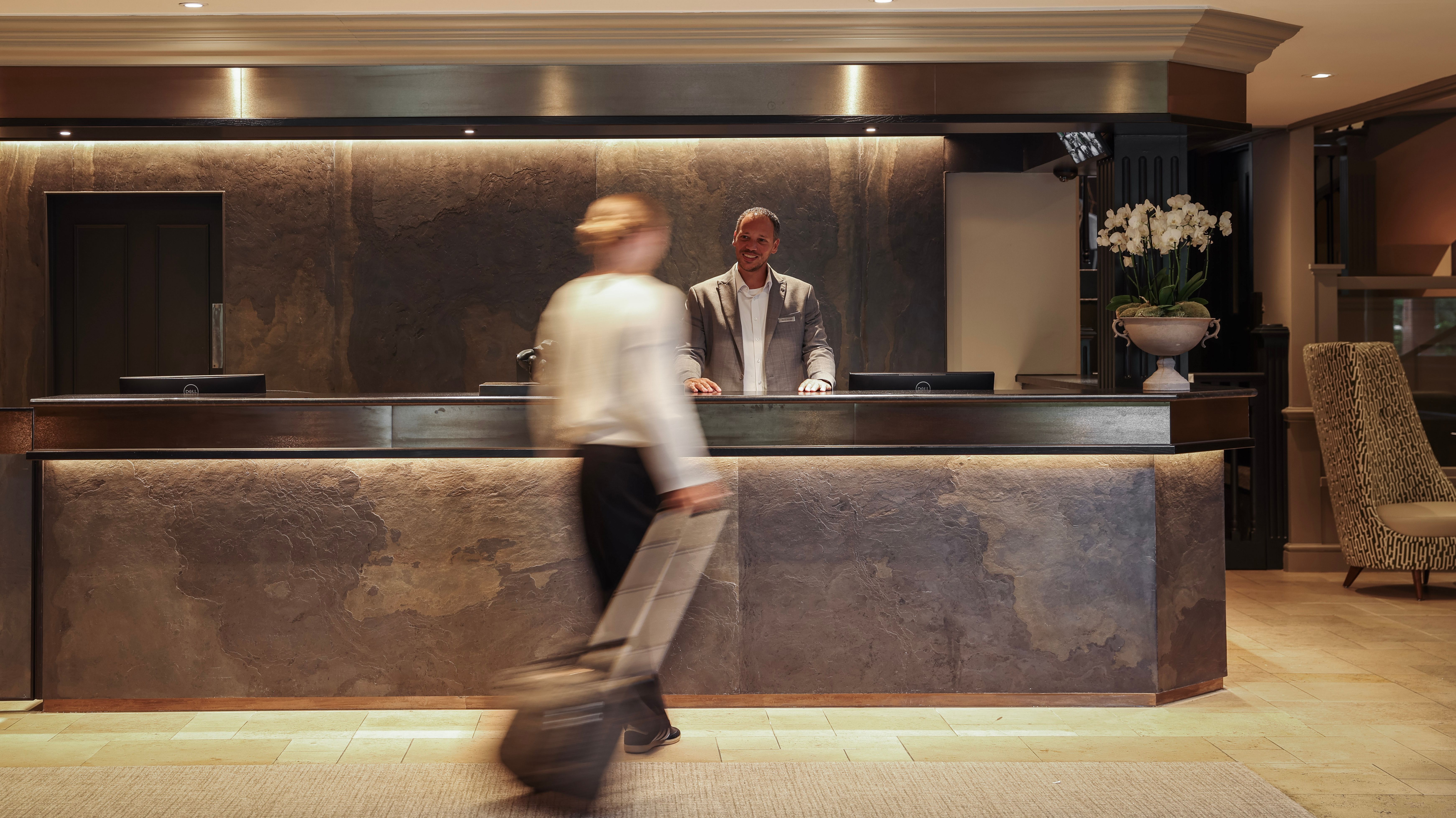 Hotel lobby reception desk with a receptionist standing behind the counter and a guest walking by with luggage.