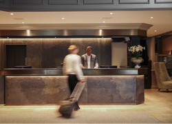 Hotel lobby reception desk with a receptionist standing behind the counter and a guest walking by with luggage.