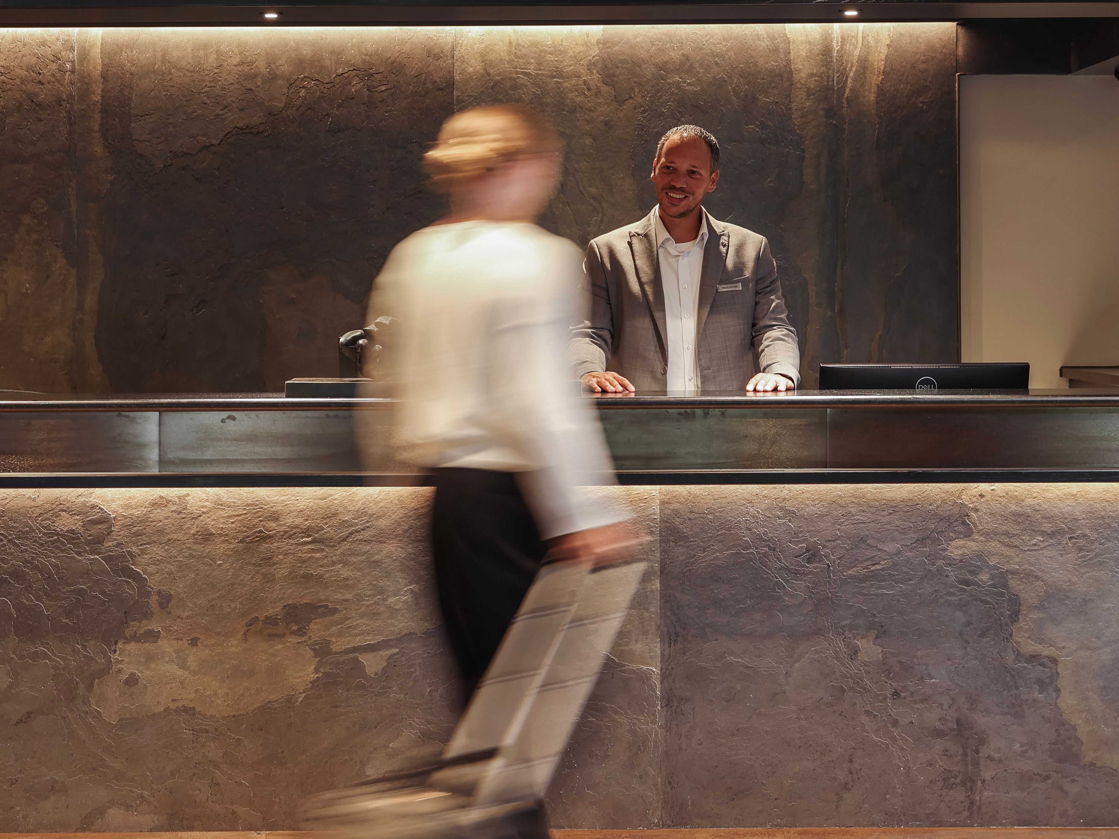 Hotel lobby reception desk with a receptionist standing behind the counter and a guest walking by with luggage.