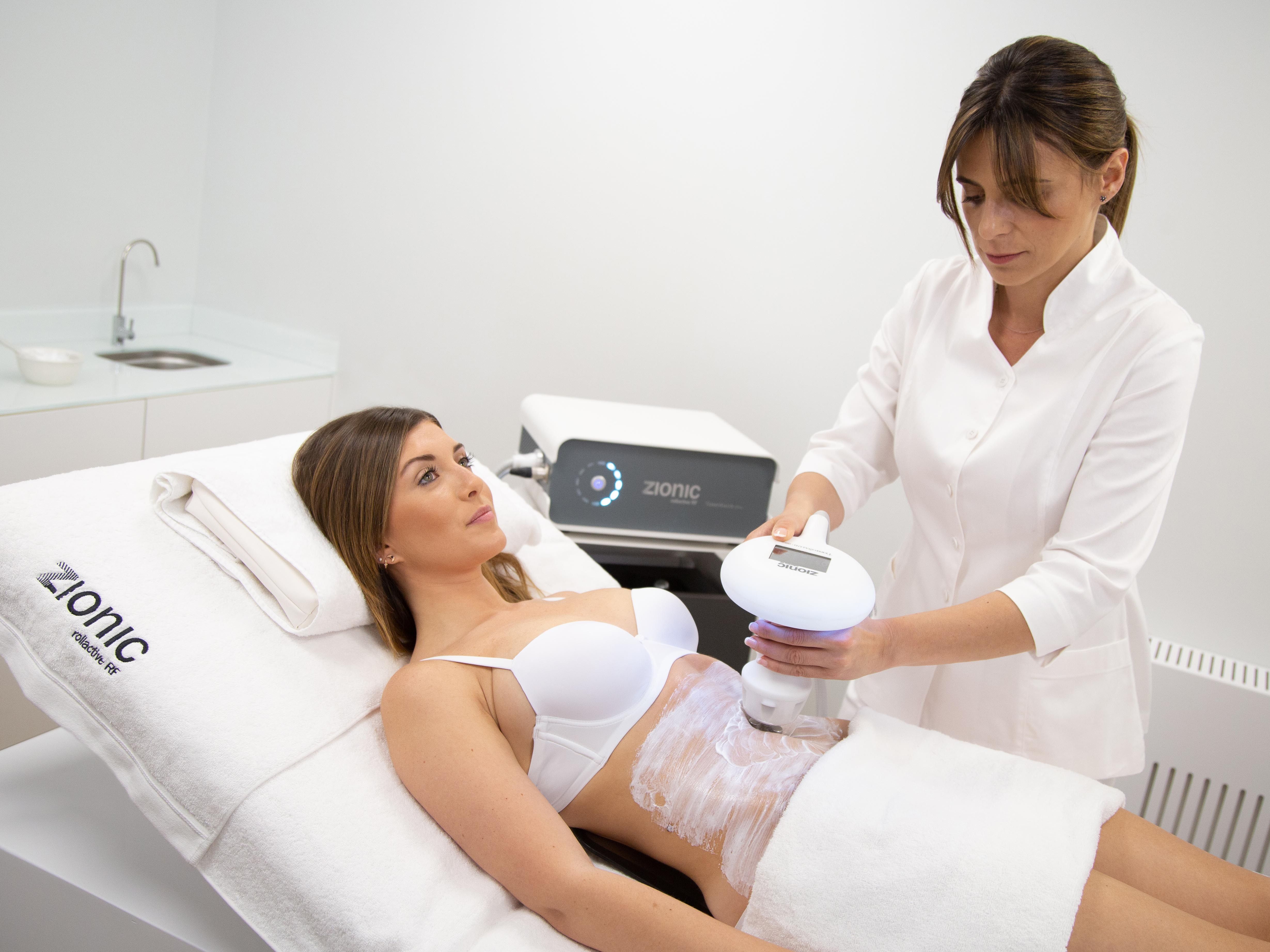 A woman receiving a body sculpting treatment using a Zionic device on her abdomen at a clinic.