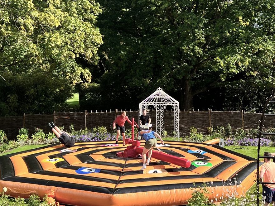 People playing on an inflatable game in a garden with large trees and a gazebo in the background.