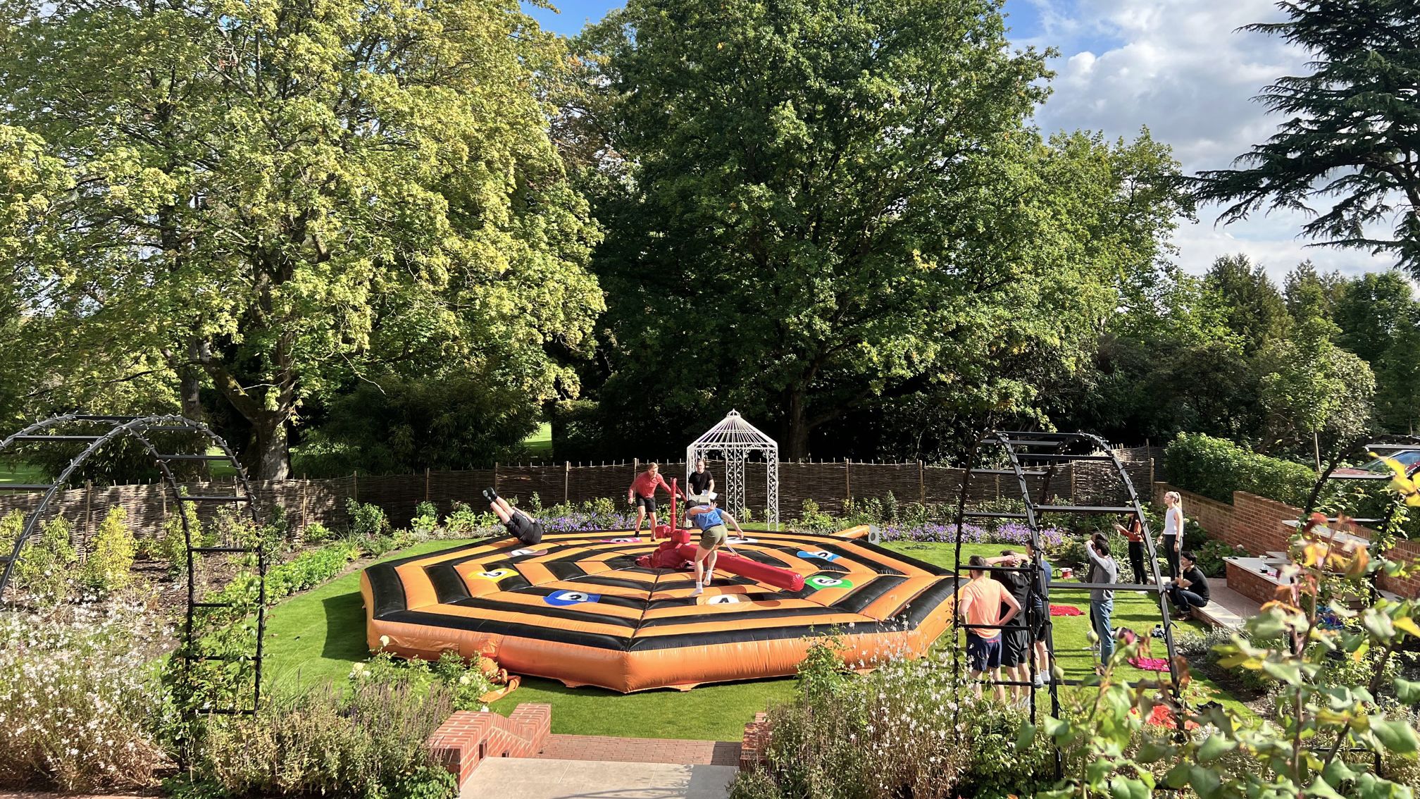 People playing on an inflatable game in a garden with large trees and a gazebo in the background.