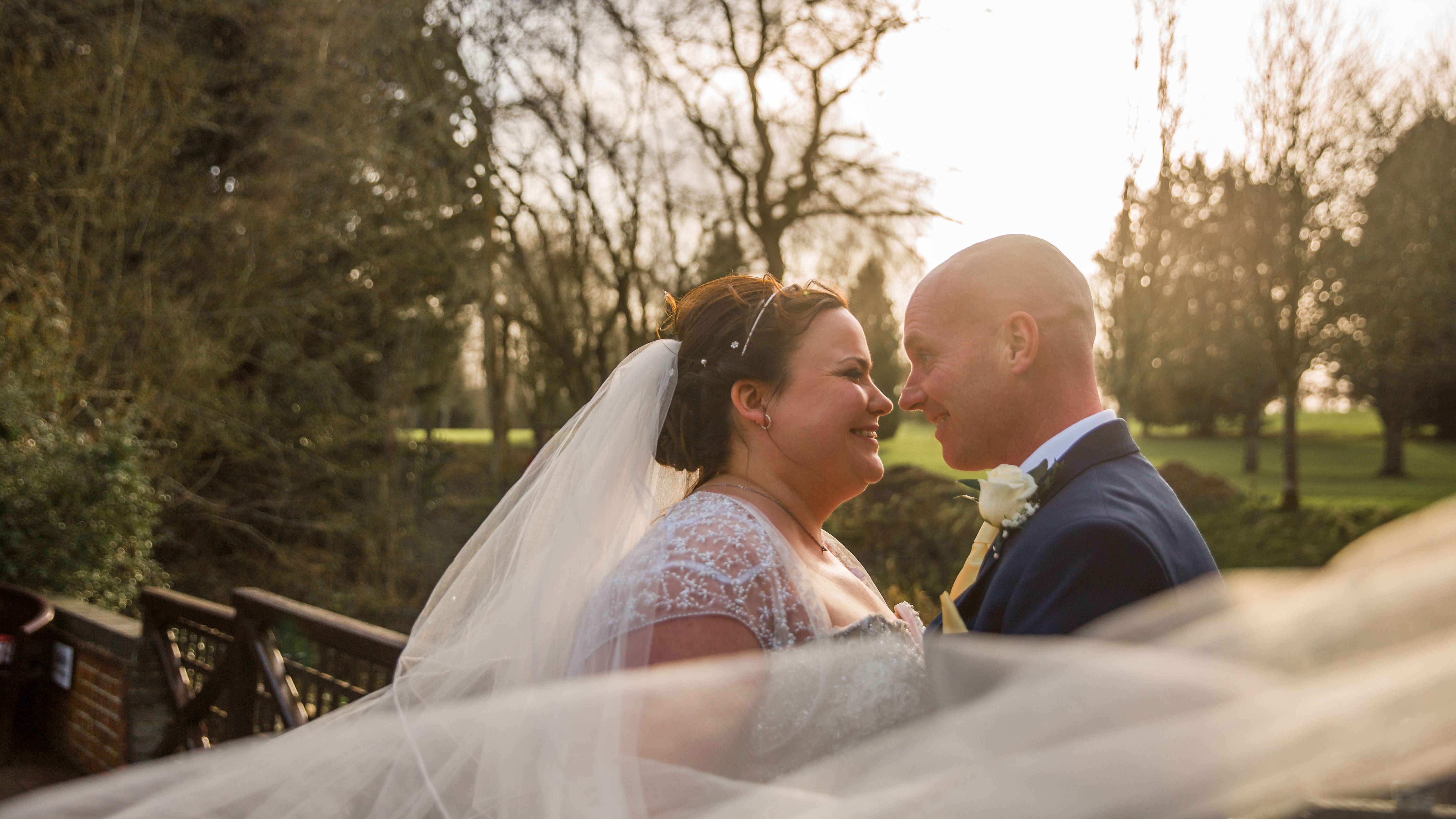 Bride and groom smiling at each other outdoors with veil blowing in the wind