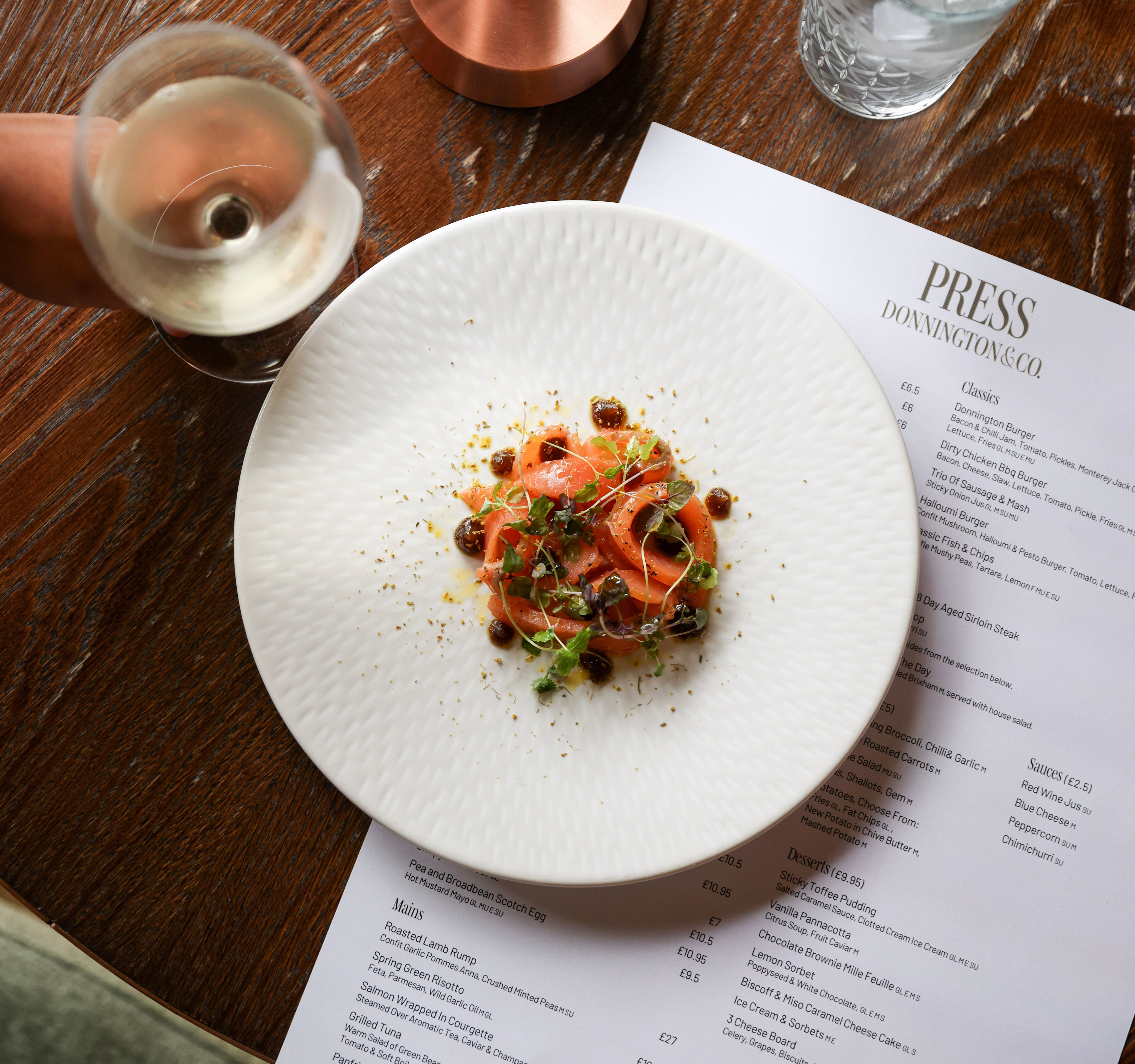 A plate of gourmet food on a wooden table next to a restaurant menu, a glass of white wine, a copper lamp, and a glass of water with ice.