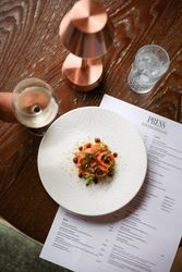 A plate of gourmet food on a wooden table next to a restaurant menu, a glass of white wine, a copper lamp, and a glass of water with ice.
