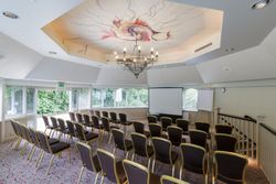 Conference room with rows of chairs facing a projector screen, decorated ceiling, and natural light from large windows.