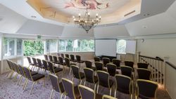 Conference room with rows of chairs facing a projector screen, decorated ceiling, and natural light from large windows.