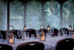 Elegant dining table set with wine glasses, lanterns with candles, and white tablecloths in a room with large windows and blinds.