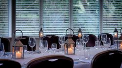 Elegant dining table set with wine glasses, lanterns with candles, and white tablecloths in a room with large windows and blinds.