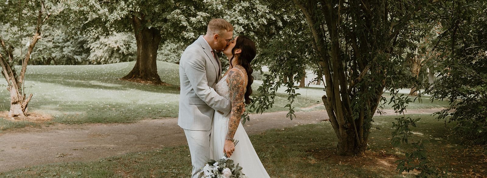 A bride and groom share a kiss outdoors under a tree, with the bride holding a bouquet and her long white dress trailing behind her.