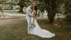 A bride and groom share a kiss outdoors under a tree, with the bride holding a bouquet and her long white dress trailing behind her.