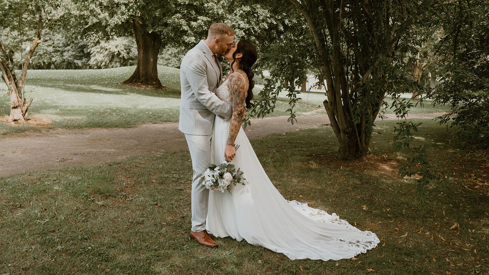 A bride and groom share a kiss outdoors under a tree, with the bride holding a bouquet and her long white dress trailing behind her.