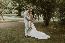 A bride and groom share a kiss outdoors under a tree, with the bride holding a bouquet and her long white dress trailing behind her.