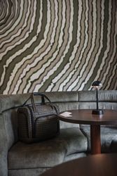 A checked travel bag on a green velvet booth seat next to a round wooden table with a small black lamp and papers.