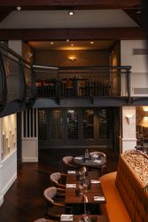 Interior of a modern restaurant with wooden tables, cushioned chairs, a wine cellar, and an upper mezzanine seating area.