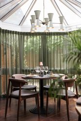 A round dining table set for four in a bright, modern sunroom with glass windows, green curtains, and a mid-century chandelier overhead.