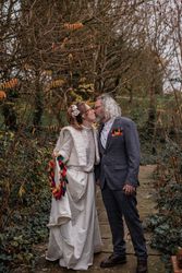 A couple in wedding attire kissing outdoors surrounded by trees and foliage.