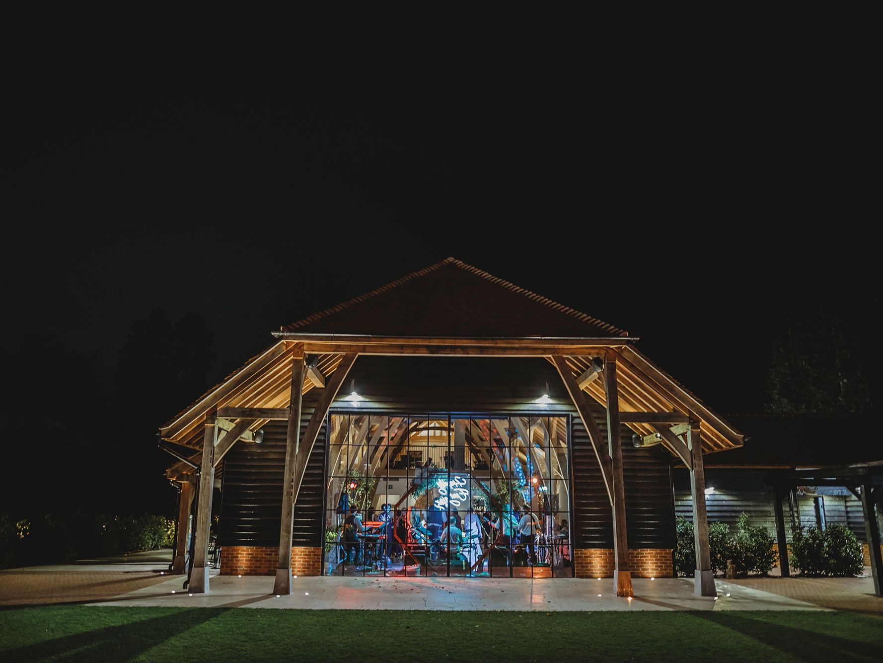 Wooden barn-style building illuminated at night with people visible inside through large glass doors.