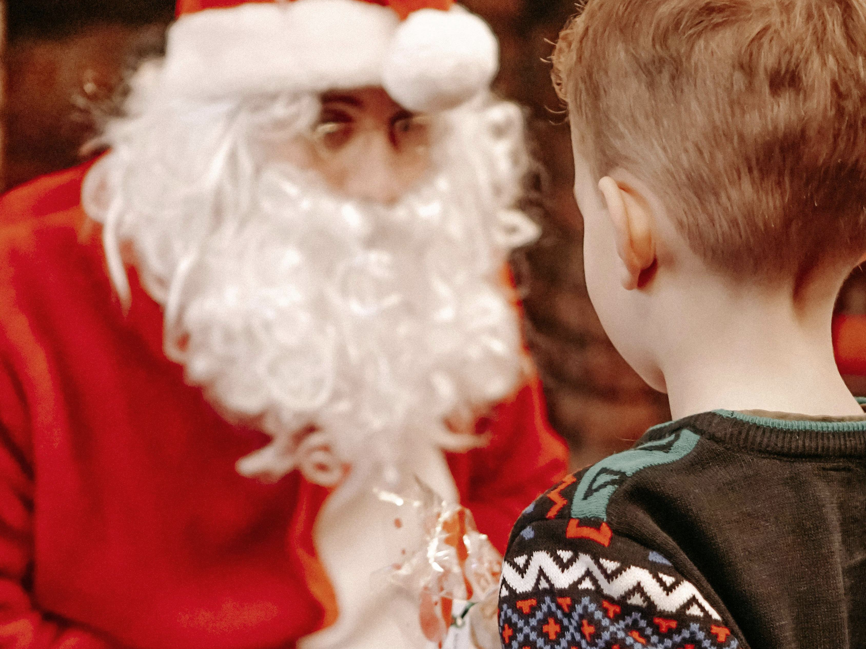 Child holding a wrapped Christmas present while standing in front of a person dressed as Santa Claus