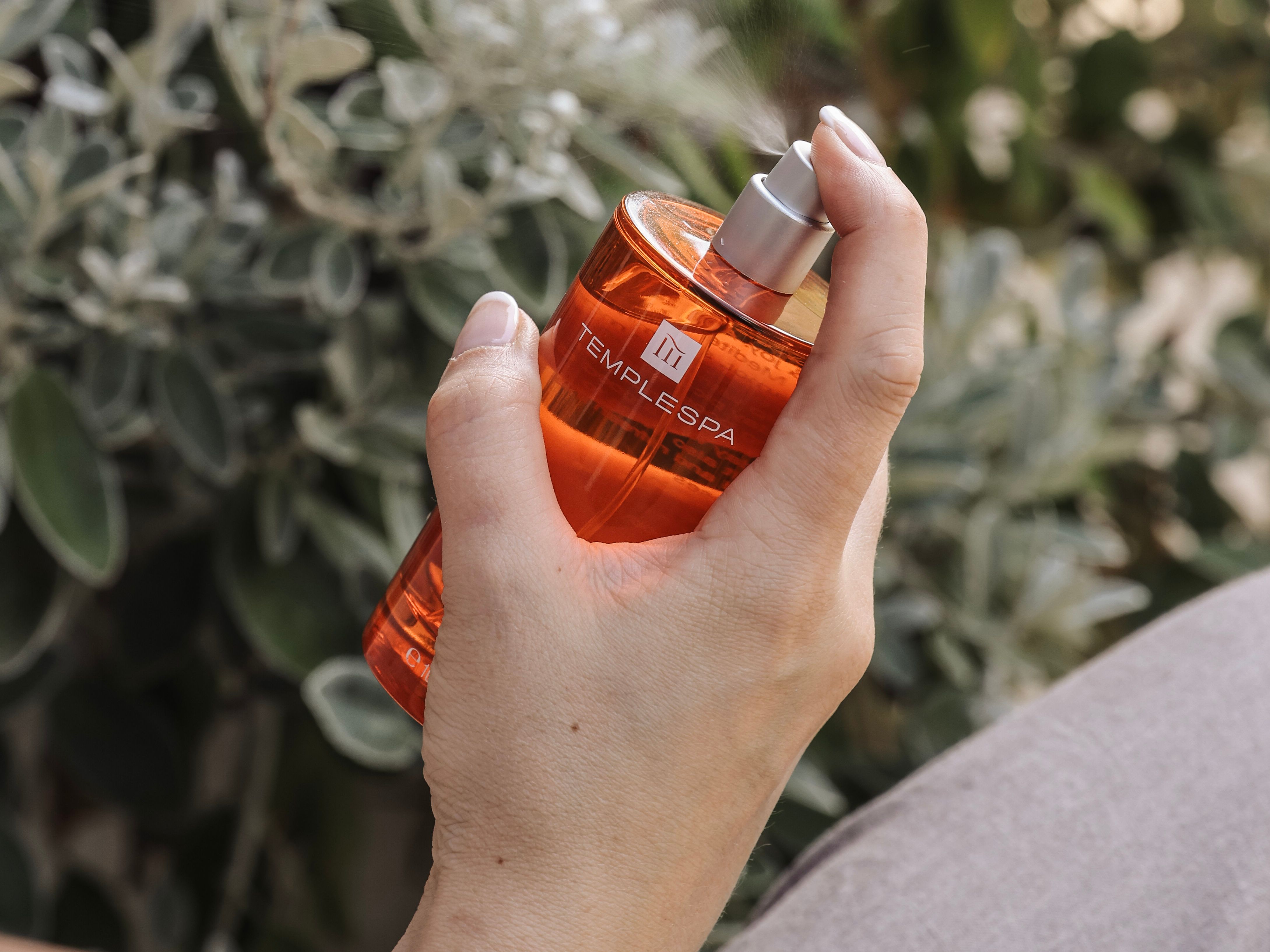 Hand holding and spraying an orange Temple Spa bottle with a leafy background