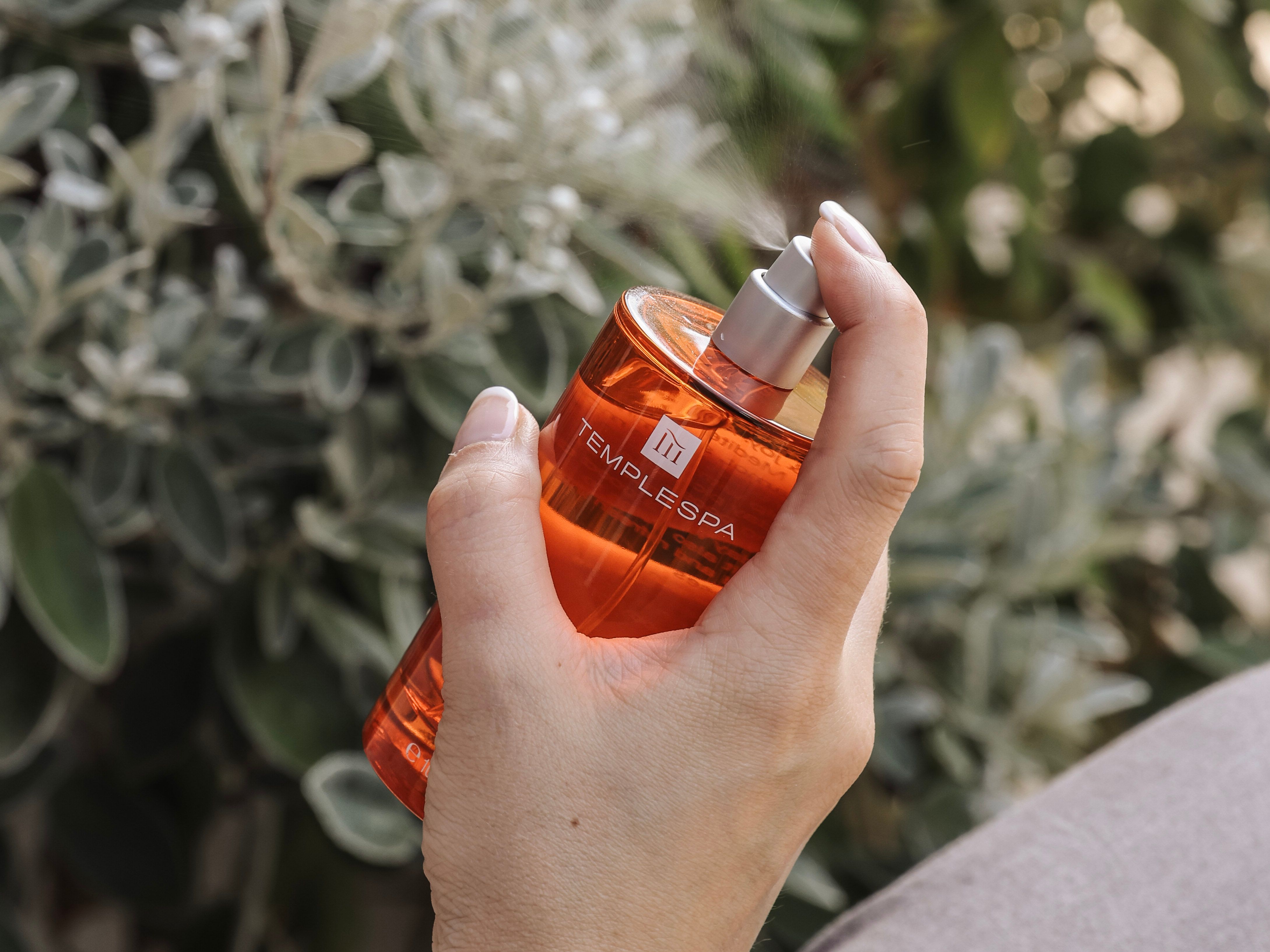 Hand holding and spraying an orange Temple Spa bottle with a leafy background