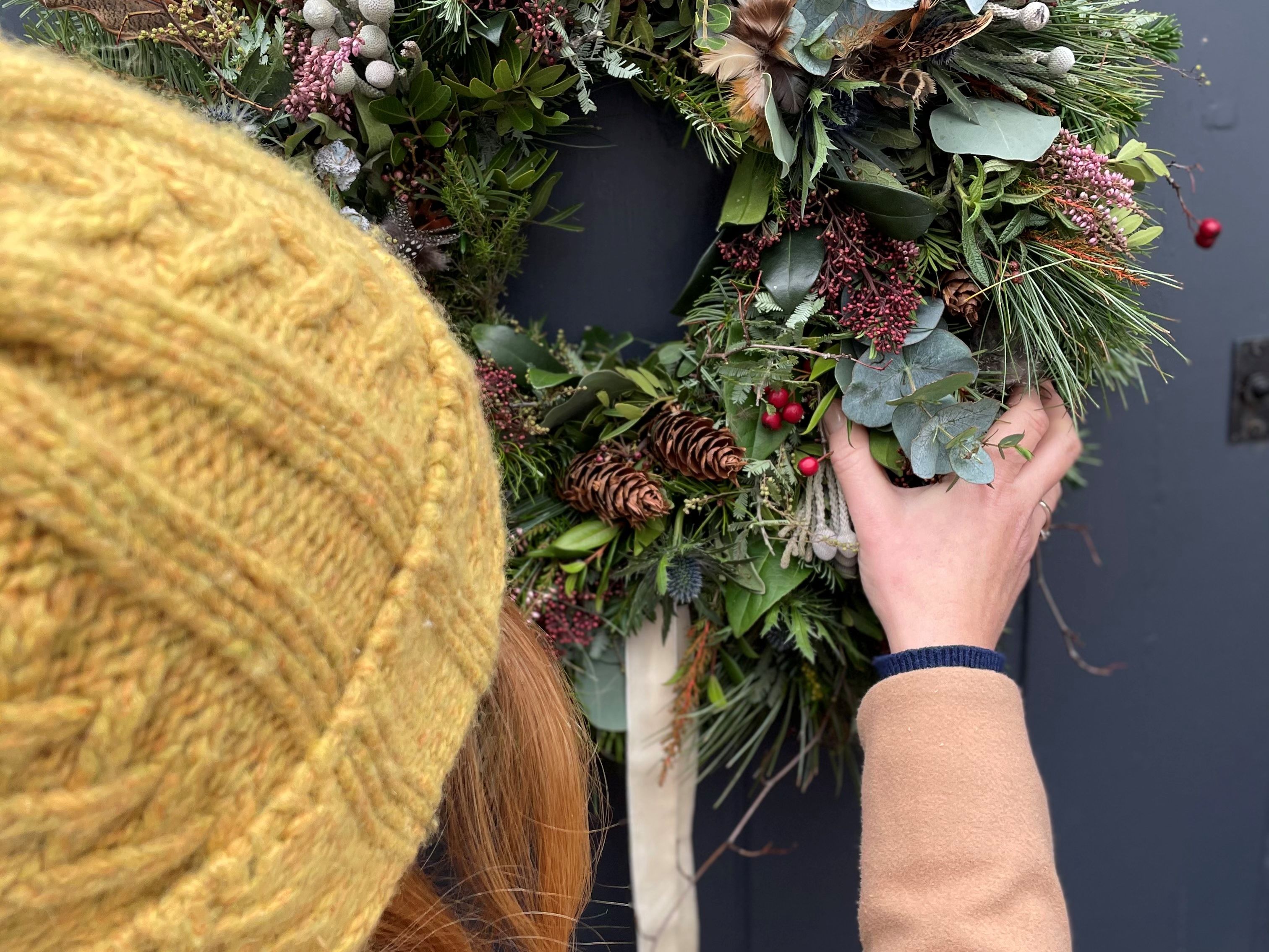 Person hanging a festive wreath decorated with pine cones, berries, and greenery on a door.
