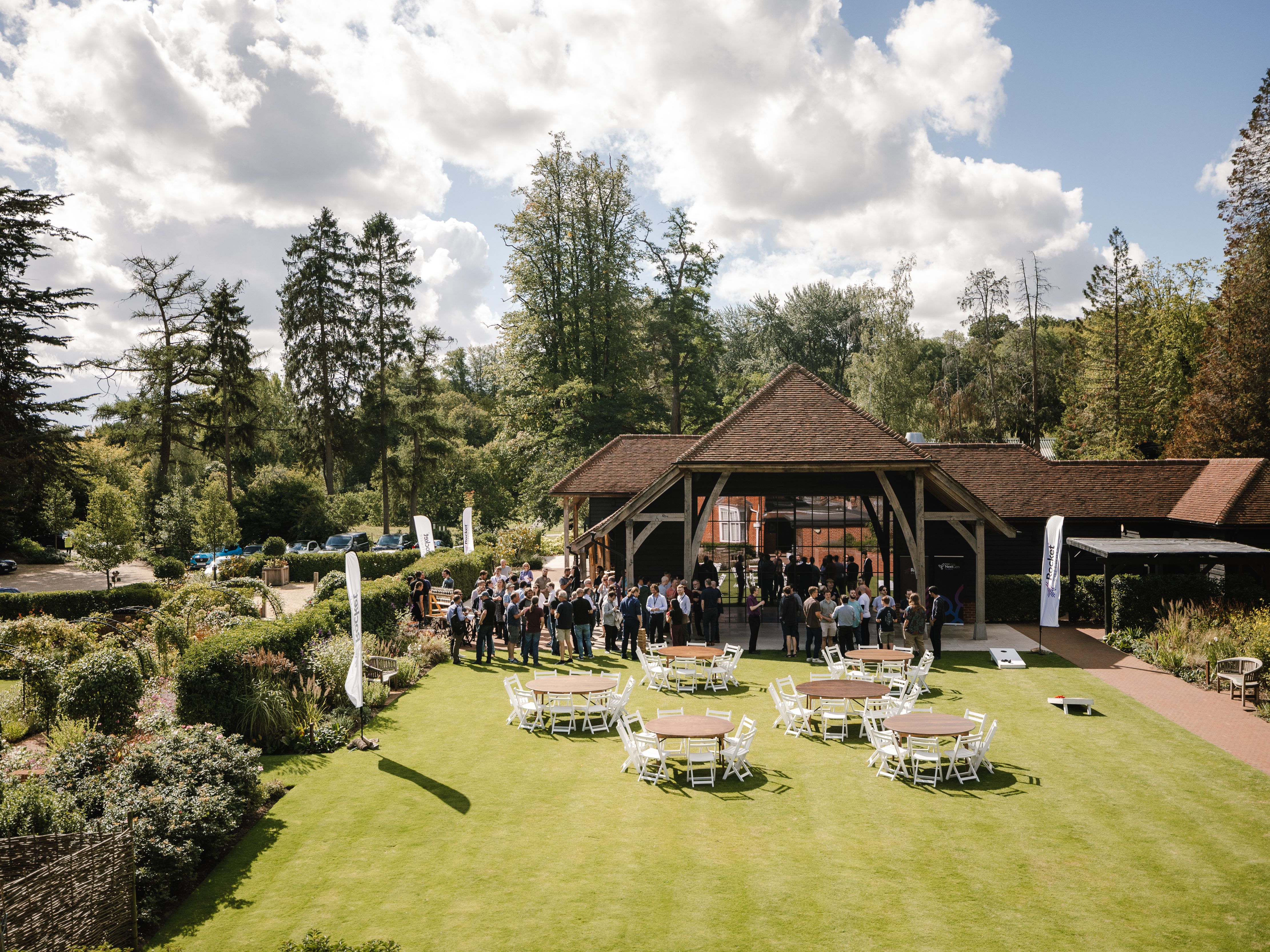Outdoor event with people gathered near a wooden pavilion, surrounded by gardens and empty round tables with chairs on a green lawn under a partly cloudy sky.