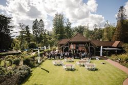 Outdoor event with people gathered near a wooden pavilion, surrounded by gardens and empty round tables with chairs on a green lawn under a partly cloudy sky.