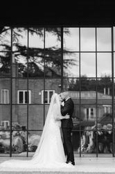 Bride and groom share a kiss in front of large glass windows reflecting trees and guests.