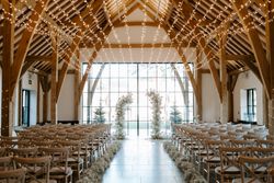 Beautifully decorated barn wedding ceremony with wooden beams, fairy lights overhead, and white floral arches at the altar.