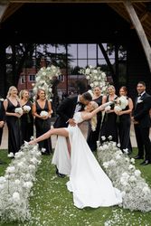 A groom dips and kisses his bride in an outdoor wedding ceremony, surrounded by bridesmaids in black dresses and groomsmen in black suits, with white floral arrangements lining the aisle.