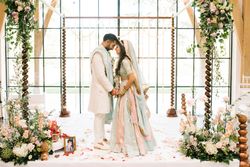 A bride and groom in traditional Indian attire stand facing each other on a flower-decorated wedding mandap.