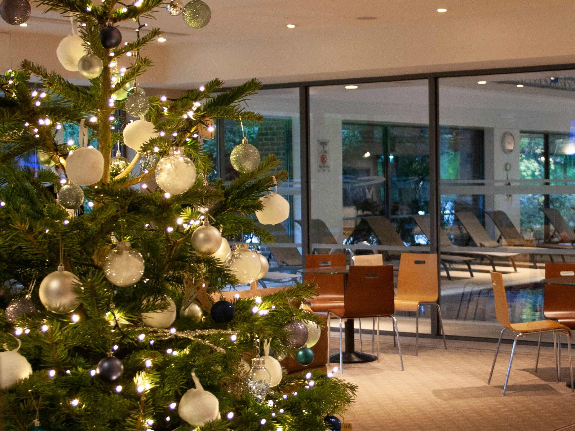 Christmas tree decorated with lights and ornaments in a spa lobby with a view of the pool area through glass walls.