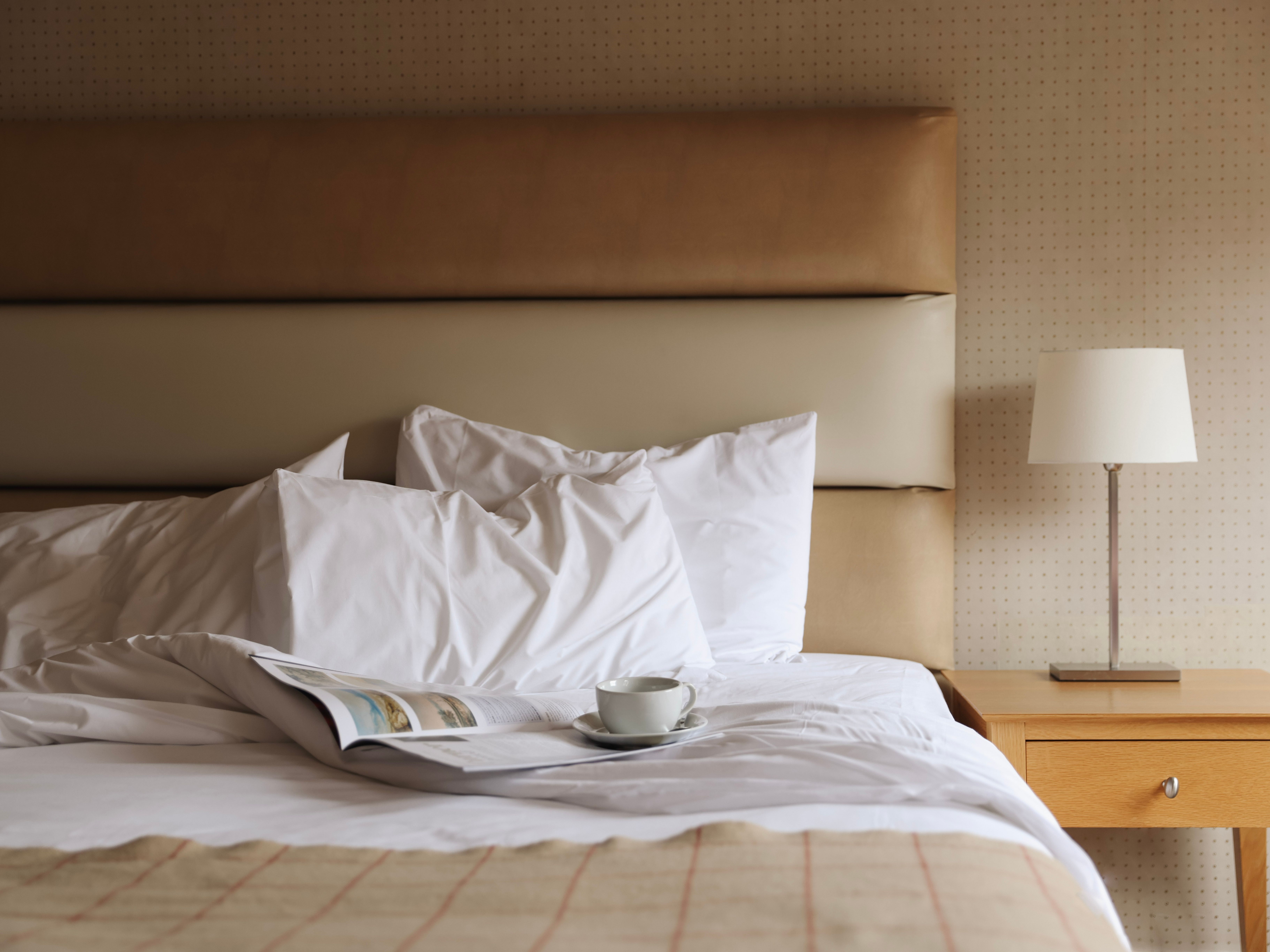 A cup and saucer on an open book resting on an unmade bed with white linens, next to a wooden bedside table and lamp.