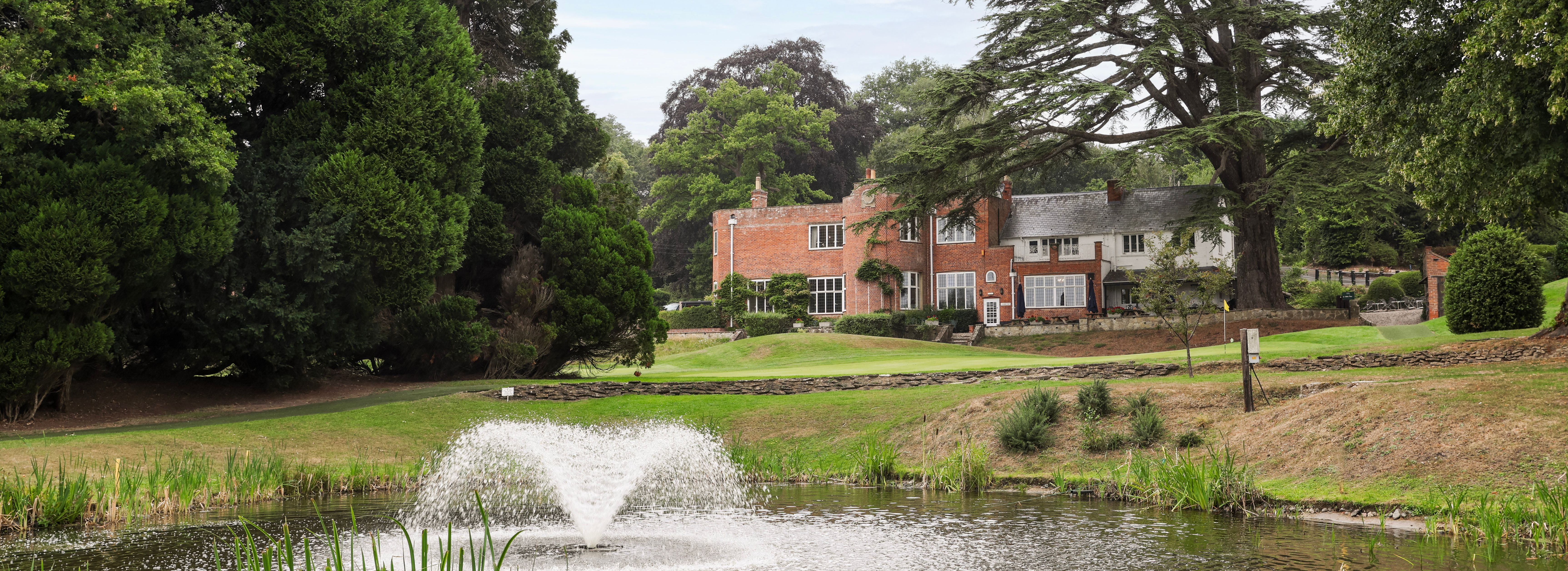 A large brick house surrounded by lush trees with a pond and water fountain in the foreground.