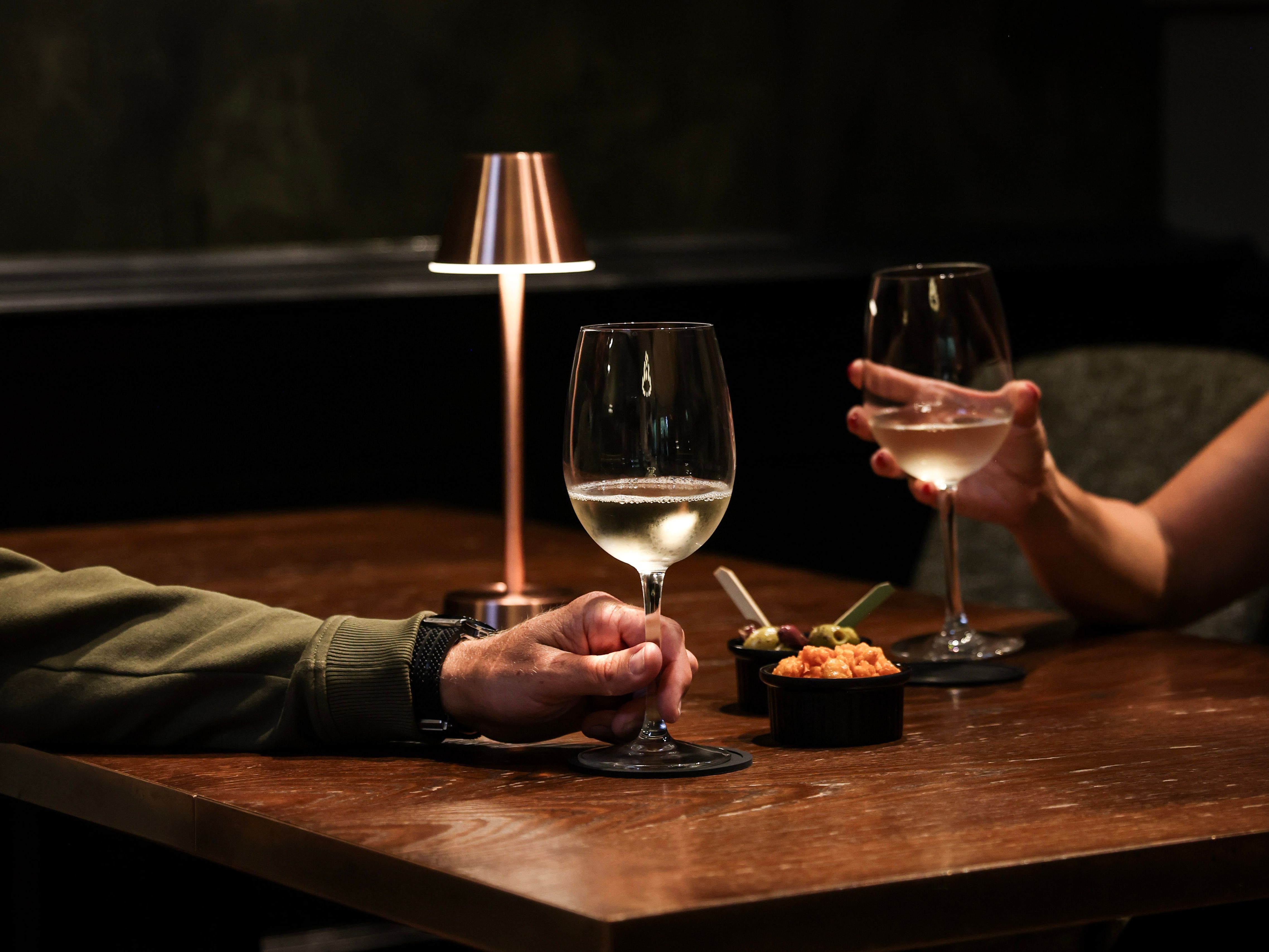 Two people clinking glasses of white wine at a dimly lit table with snacks and a small lamp.