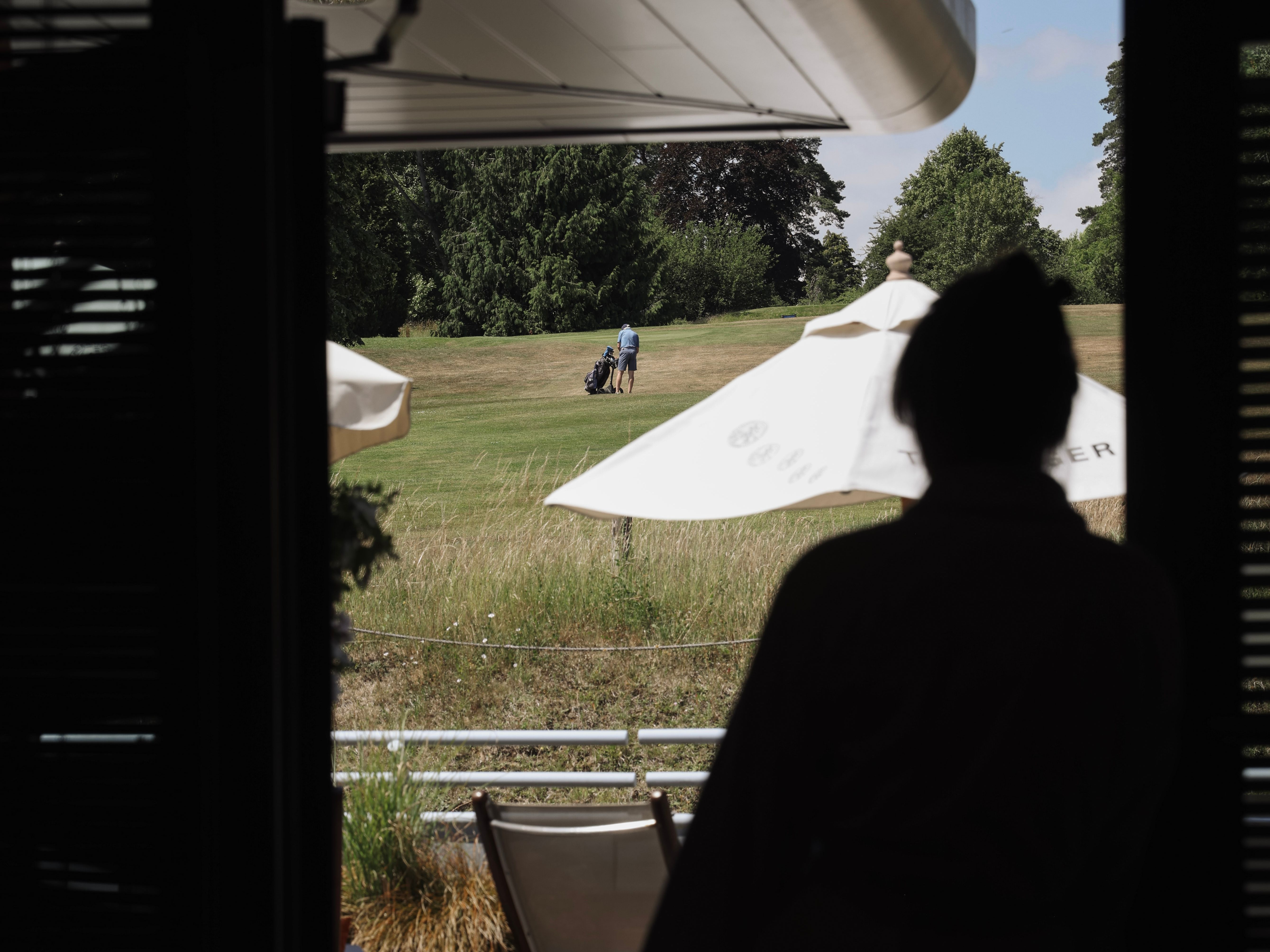 A person silhouetted in a doorway looking out at a grassy field where two people are walking with a golf bag.