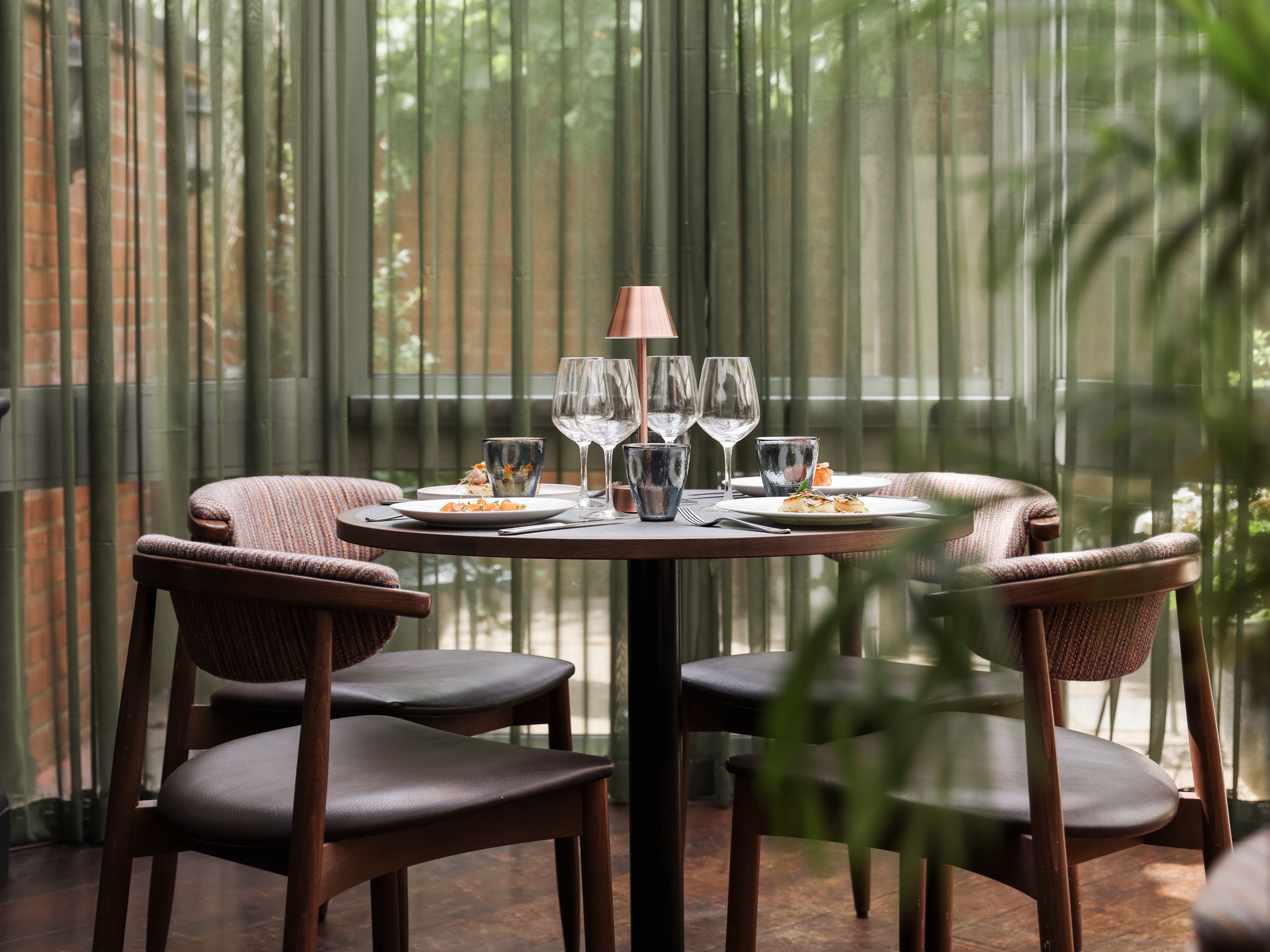 A cozy dining table set for four inside a sunlit conservatory with green curtains and a modern chandelier overhead.