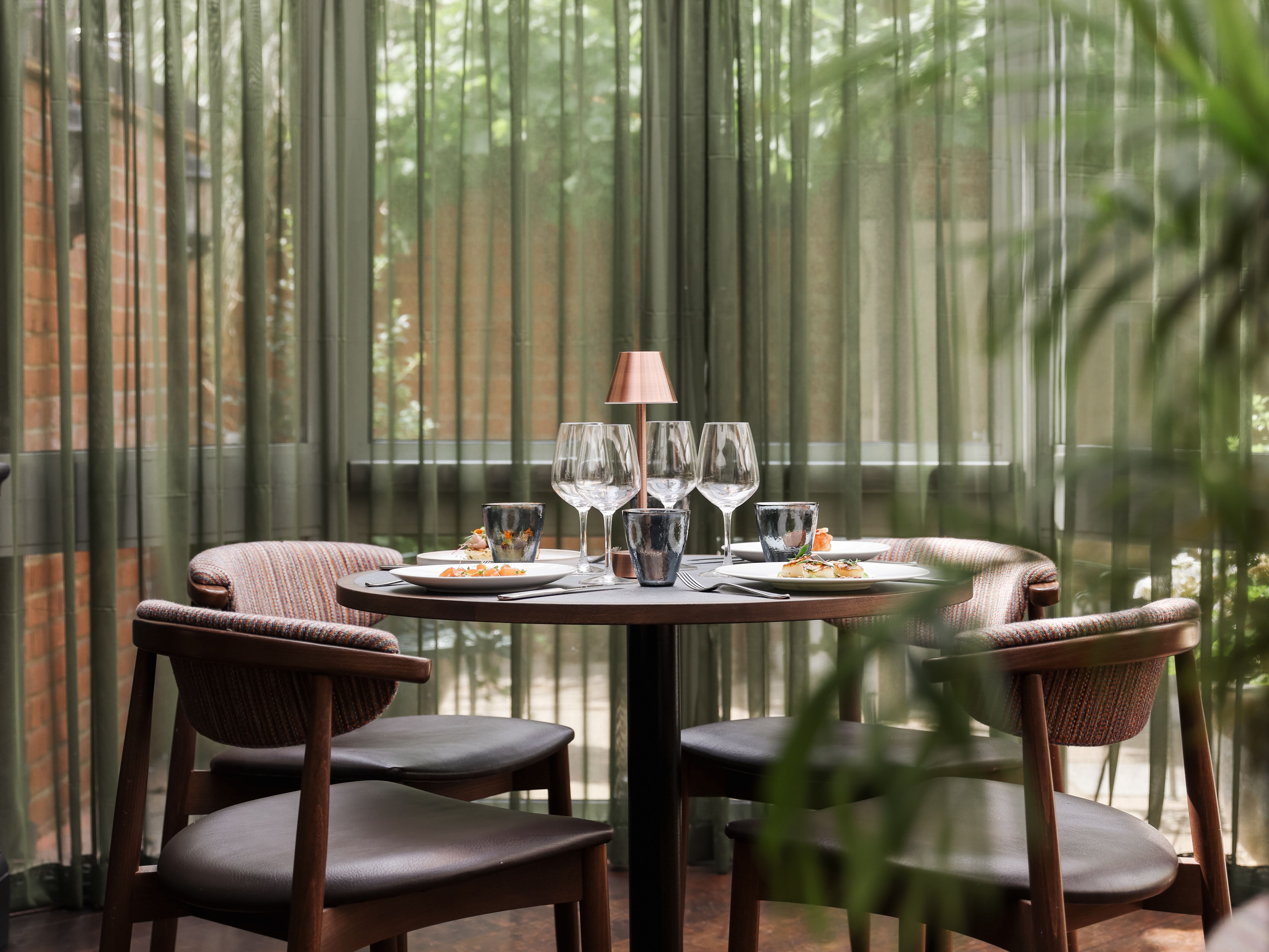 A cozy dining table set for four inside a sunlit conservatory with green curtains and a modern chandelier overhead.