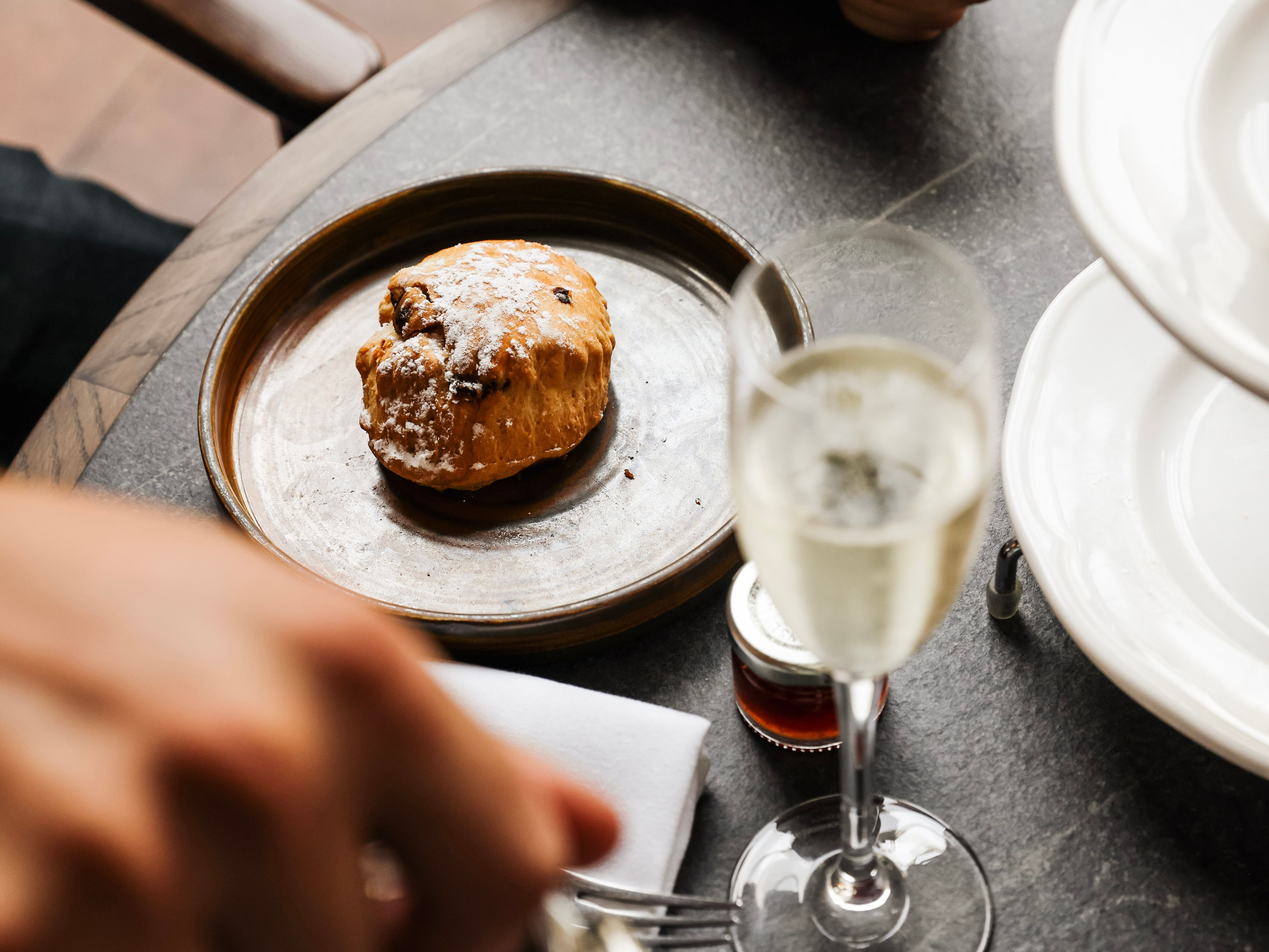 A close-up of a scone on a plate, a glass of champagne, and a hand pouring tea at a table set for afternoon tea.