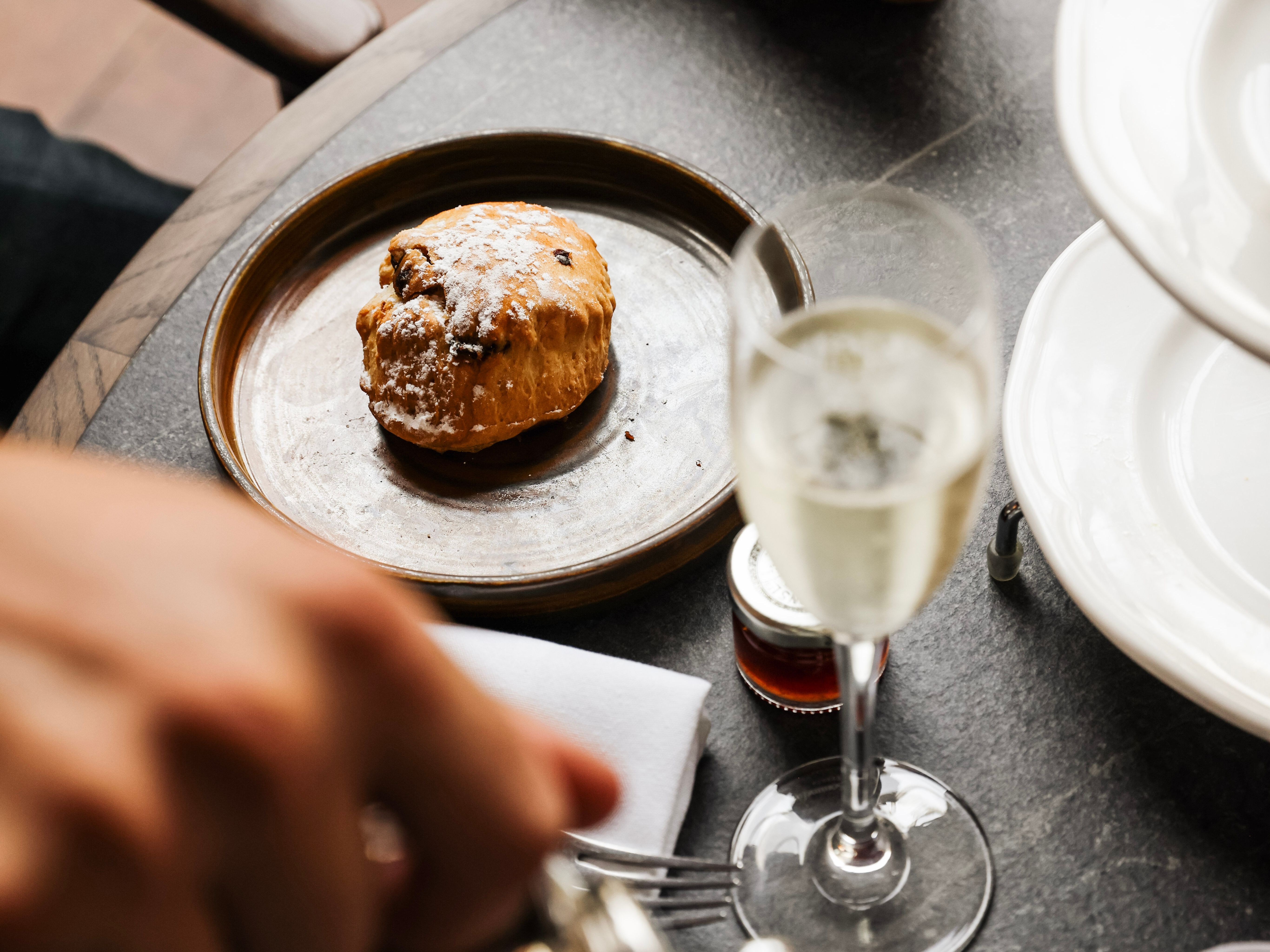 A close-up of a scone on a plate, a glass of champagne, and a hand pouring tea at a table set for afternoon tea.