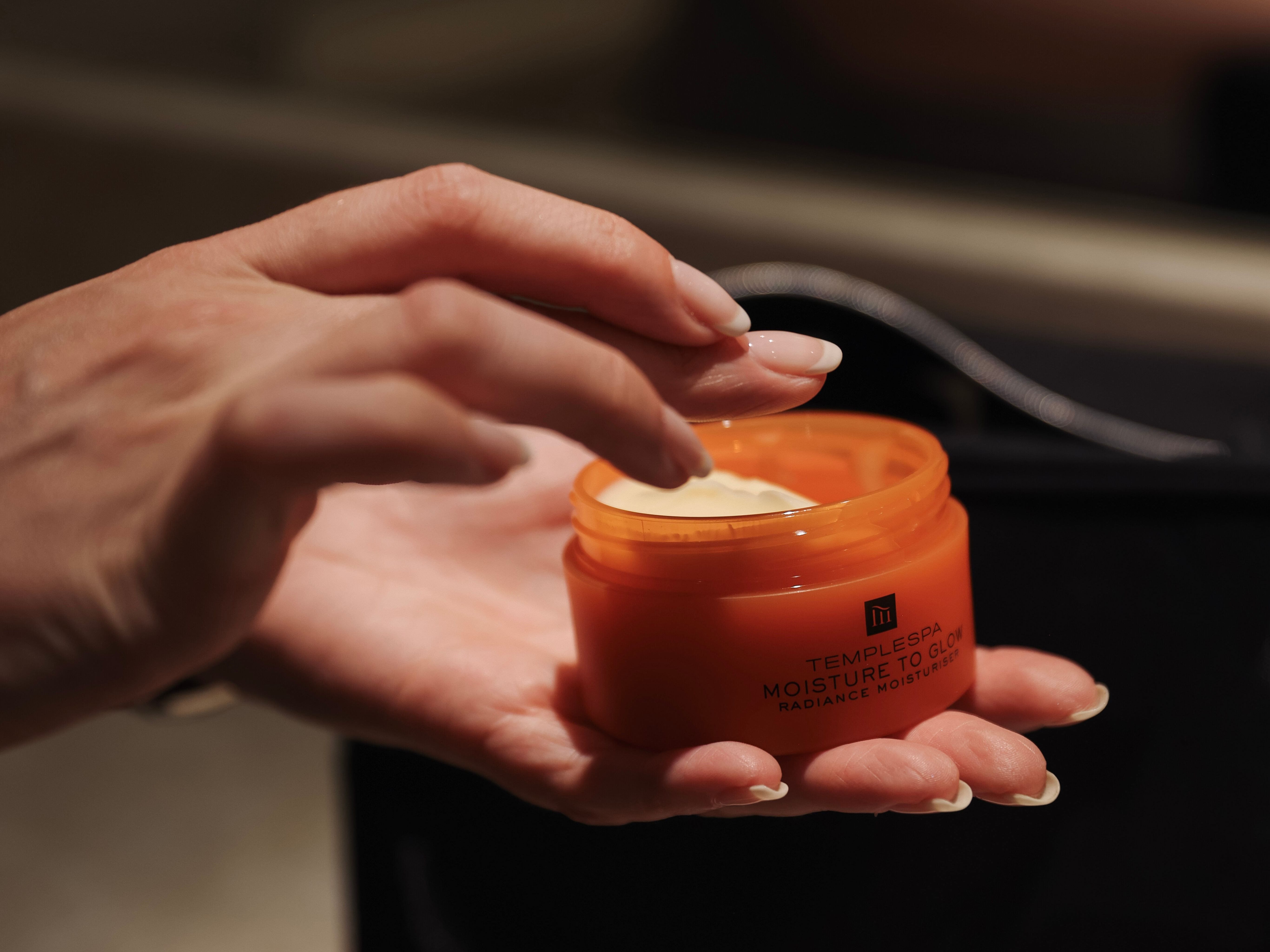 Hand holding an orange jar of Temple Spa Moisture To Glow Radiance Moisturiser, with a finger scooping out cream.