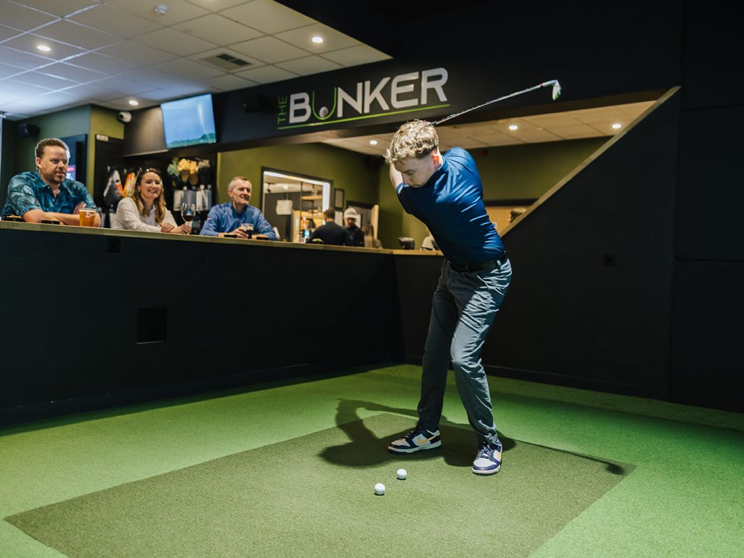 Man swinging a golf club indoors with several people watching from behind a counter.