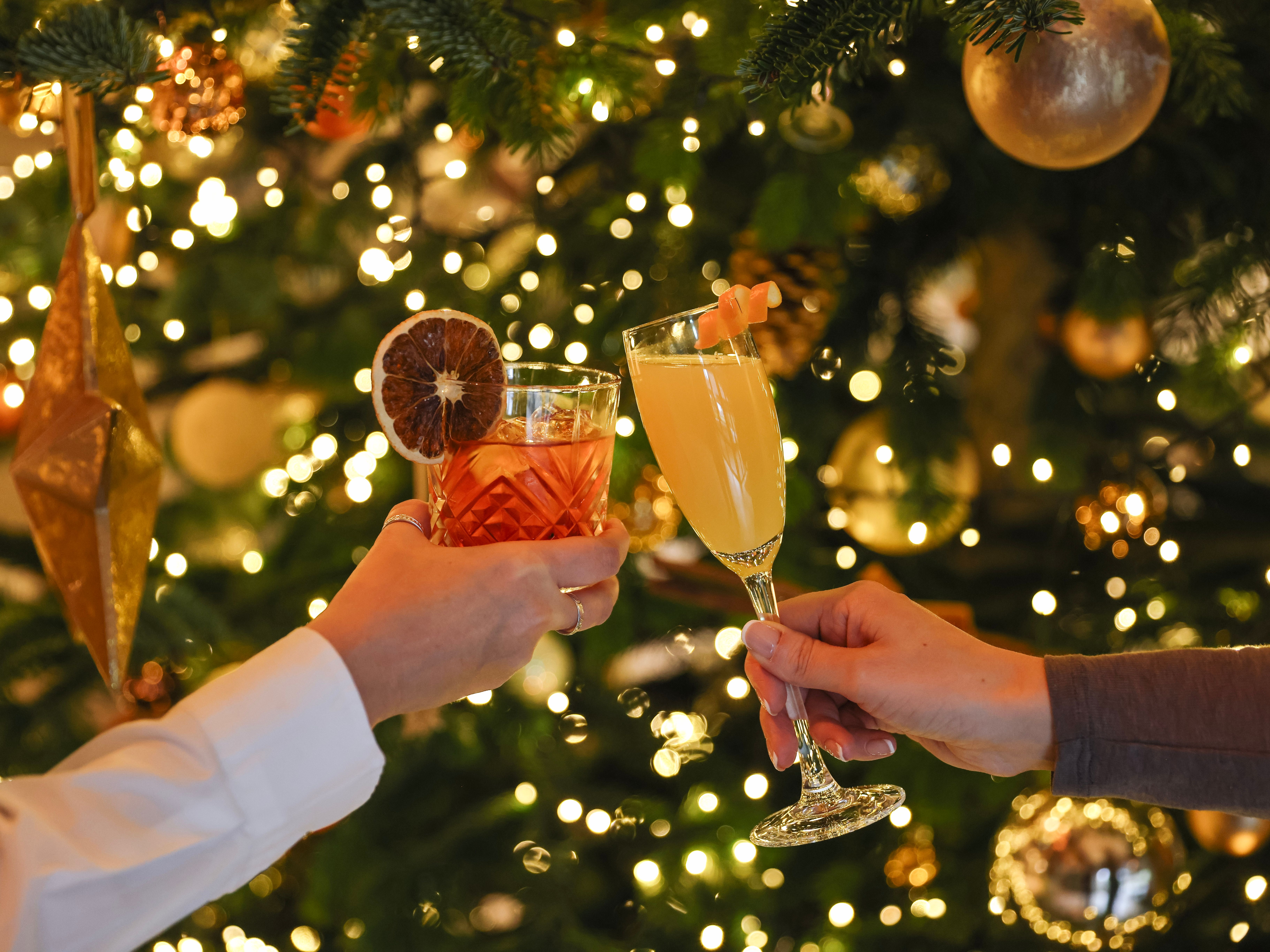 Two people clinking festive cocktails in front of a decorated Christmas tree with golden ornaments and lights.