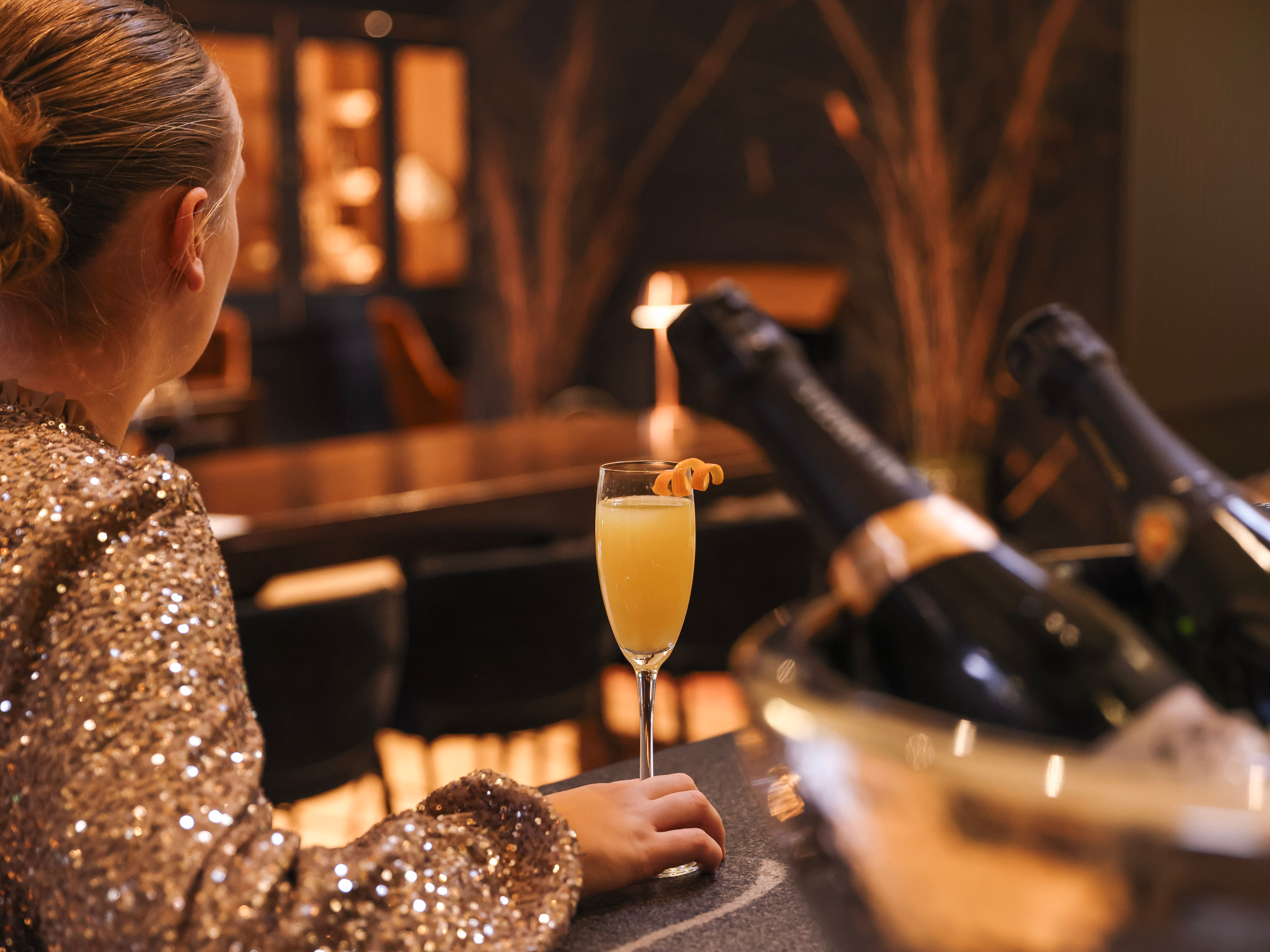 Person in a sparkly outfit sitting at a bar with a glass of orange cocktail and bottles of champagne in an ice bucket.