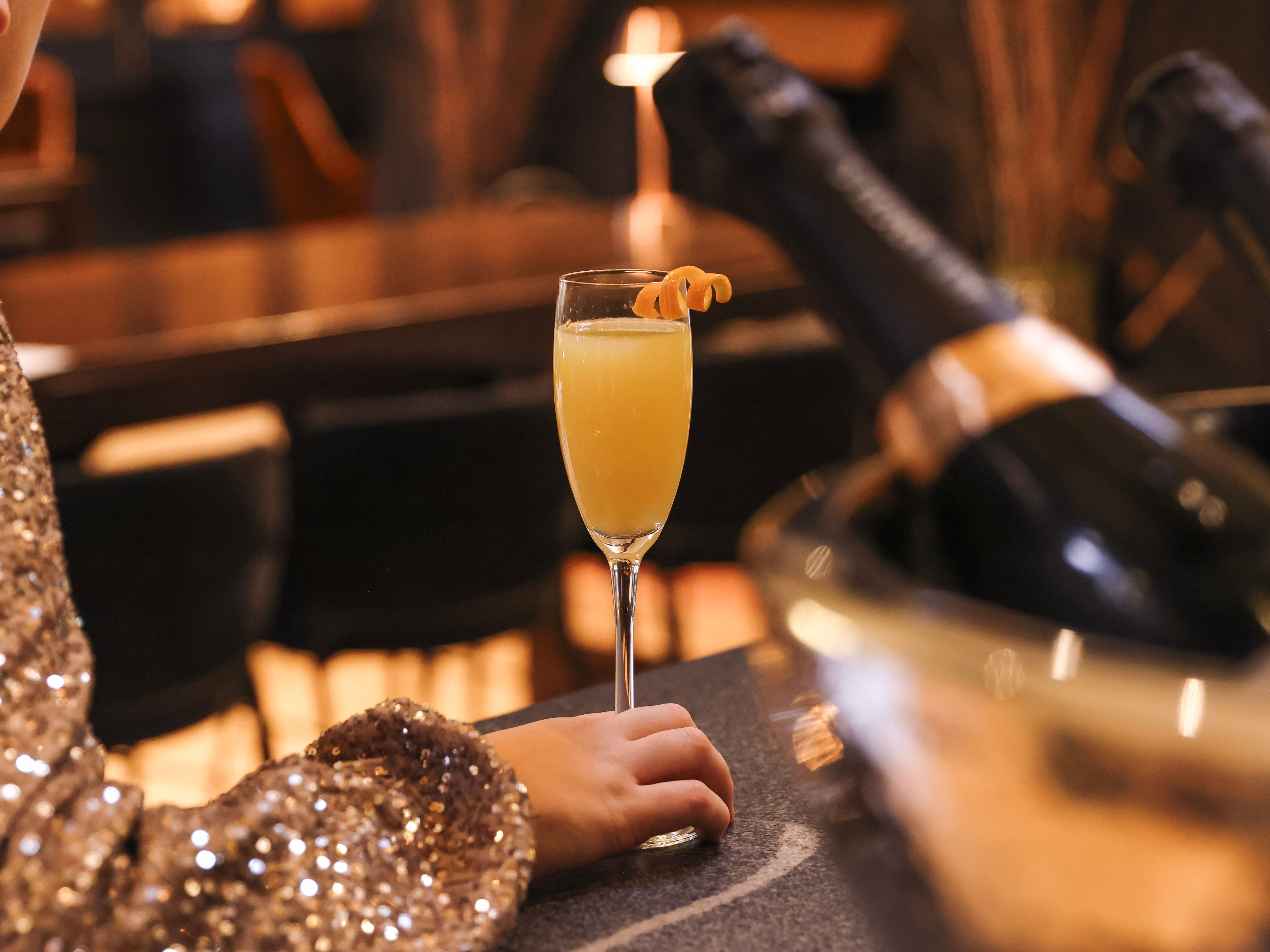 Person in a sparkly outfit sitting at a bar with a glass of orange cocktail and bottles of champagne in an ice bucket.