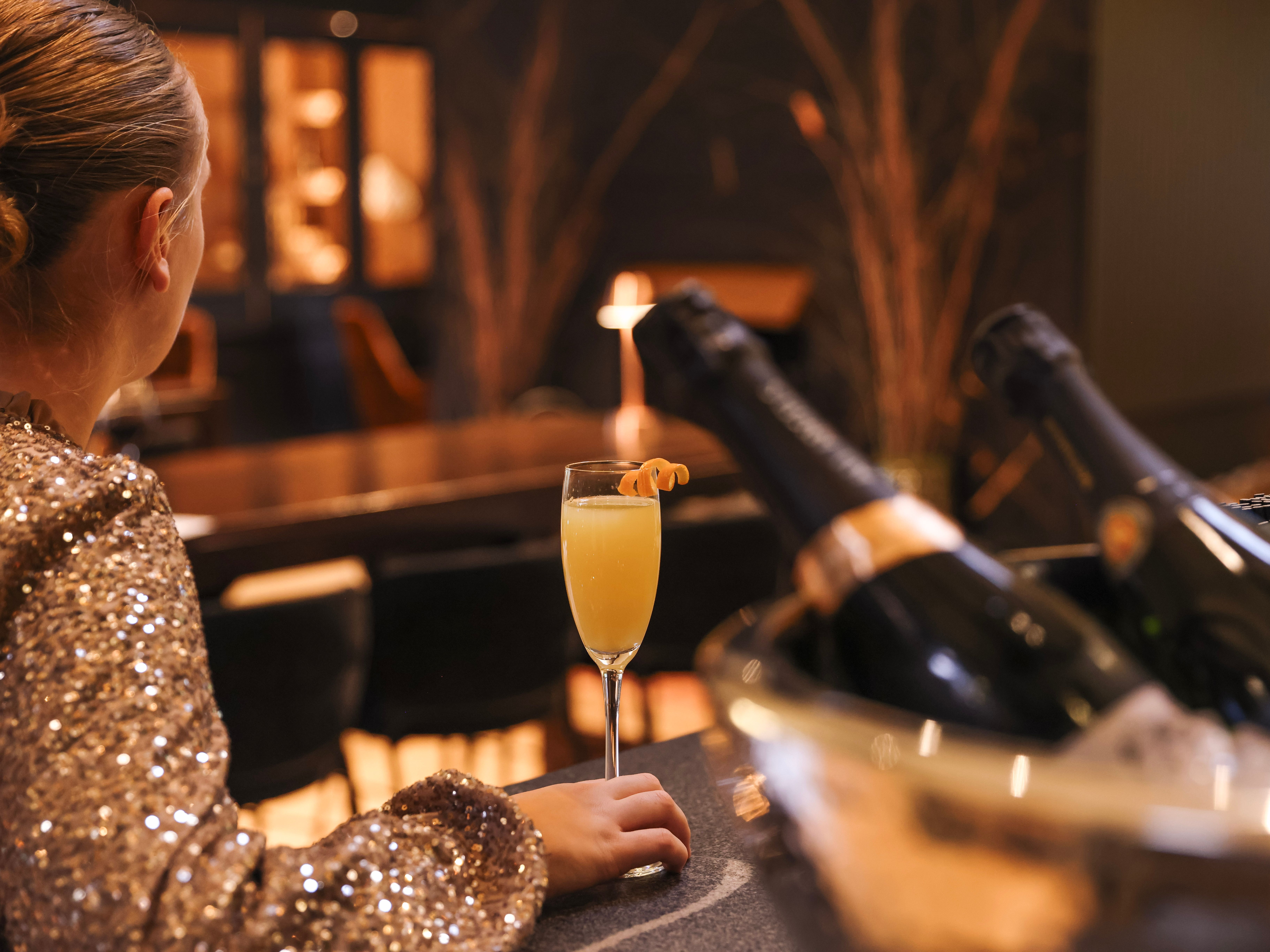 Person in a sparkly outfit sitting at a bar with a glass of orange cocktail and bottles of champagne in an ice bucket.
