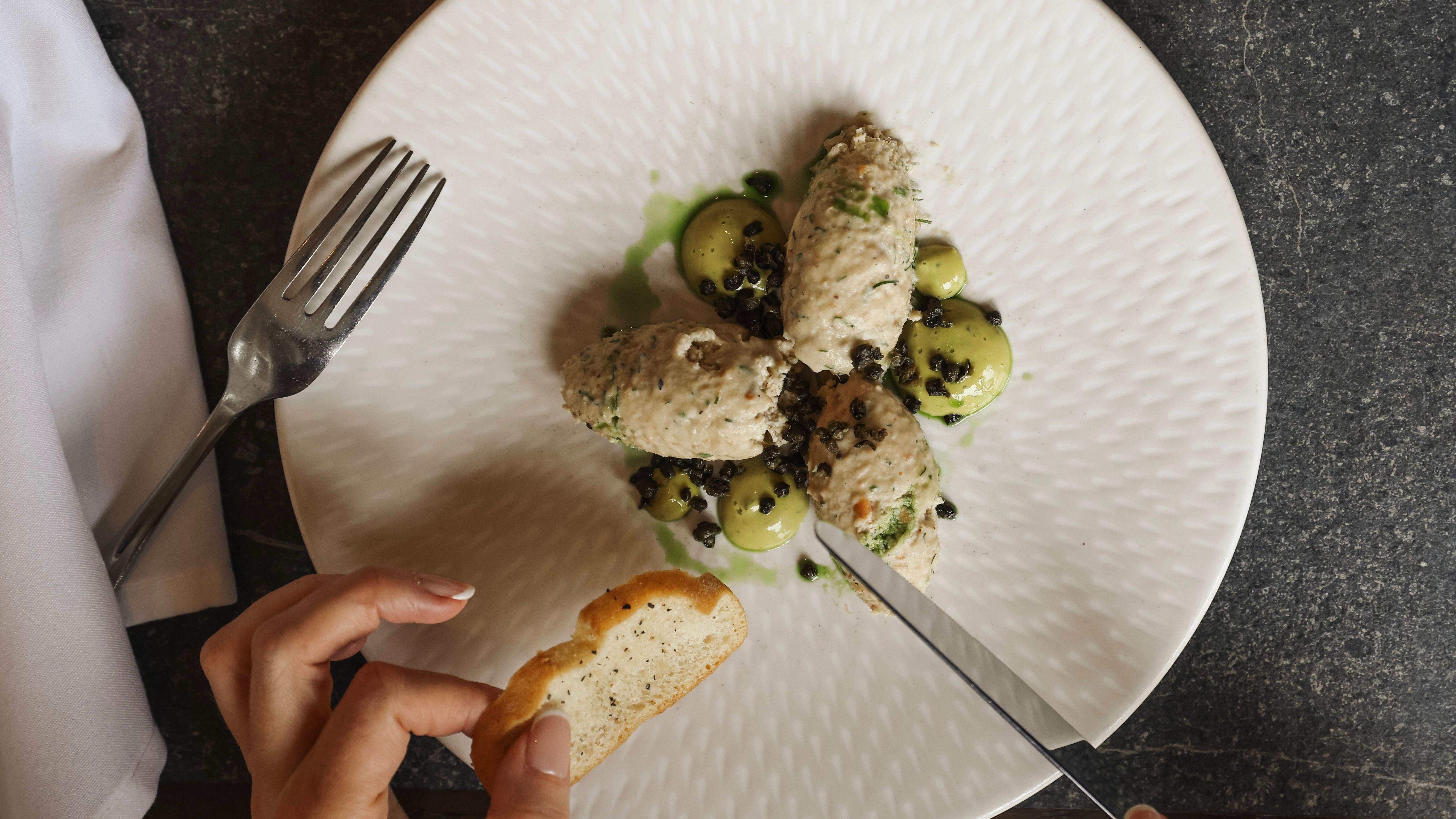A person holding a slice of toasted bread while cutting into a plated gourmet dish, accompanied by a fork and a glass of white wine