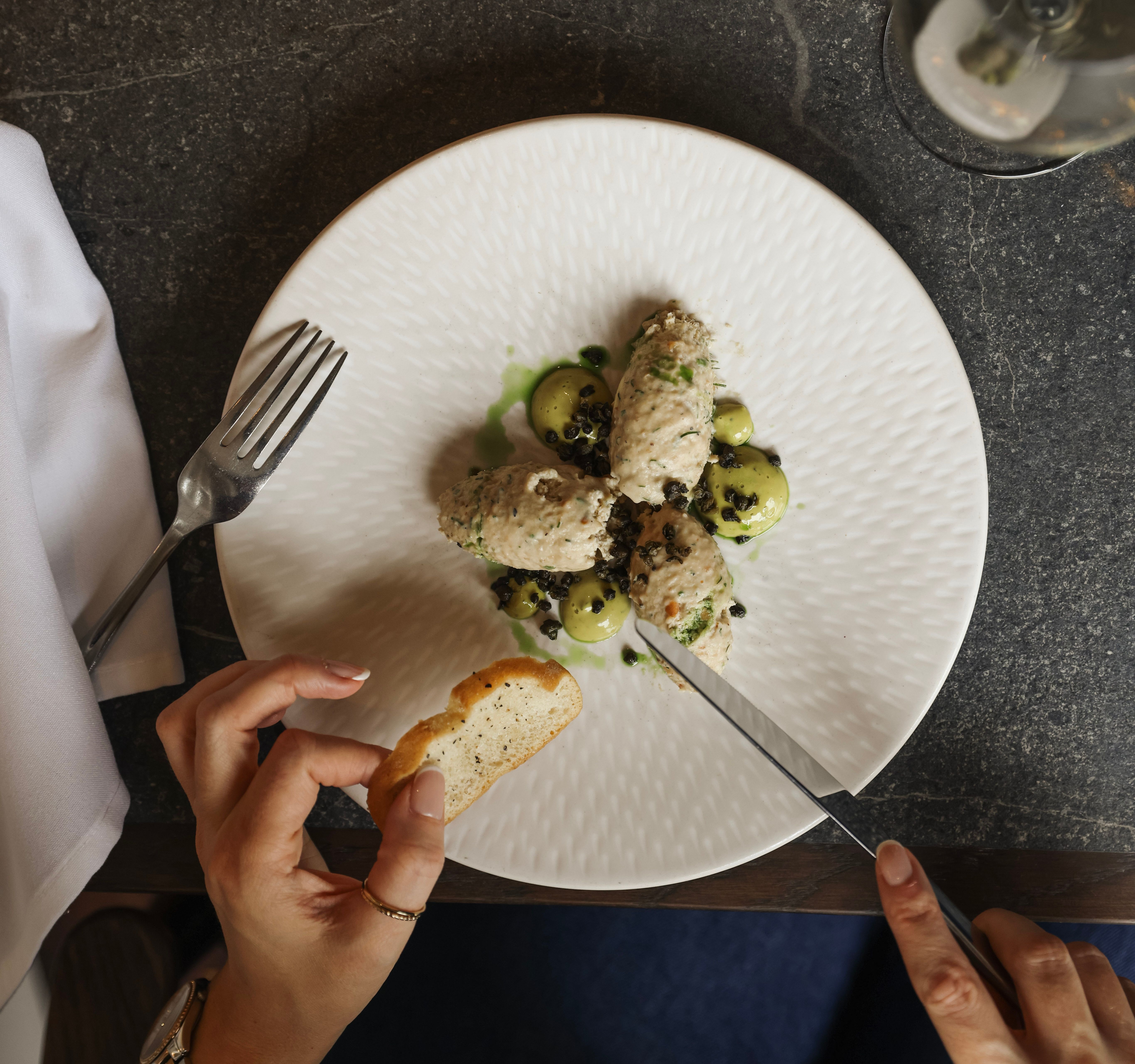 A person holding a slice of toasted bread while cutting into a plated gourmet dish, accompanied by a fork and a glass of white wine