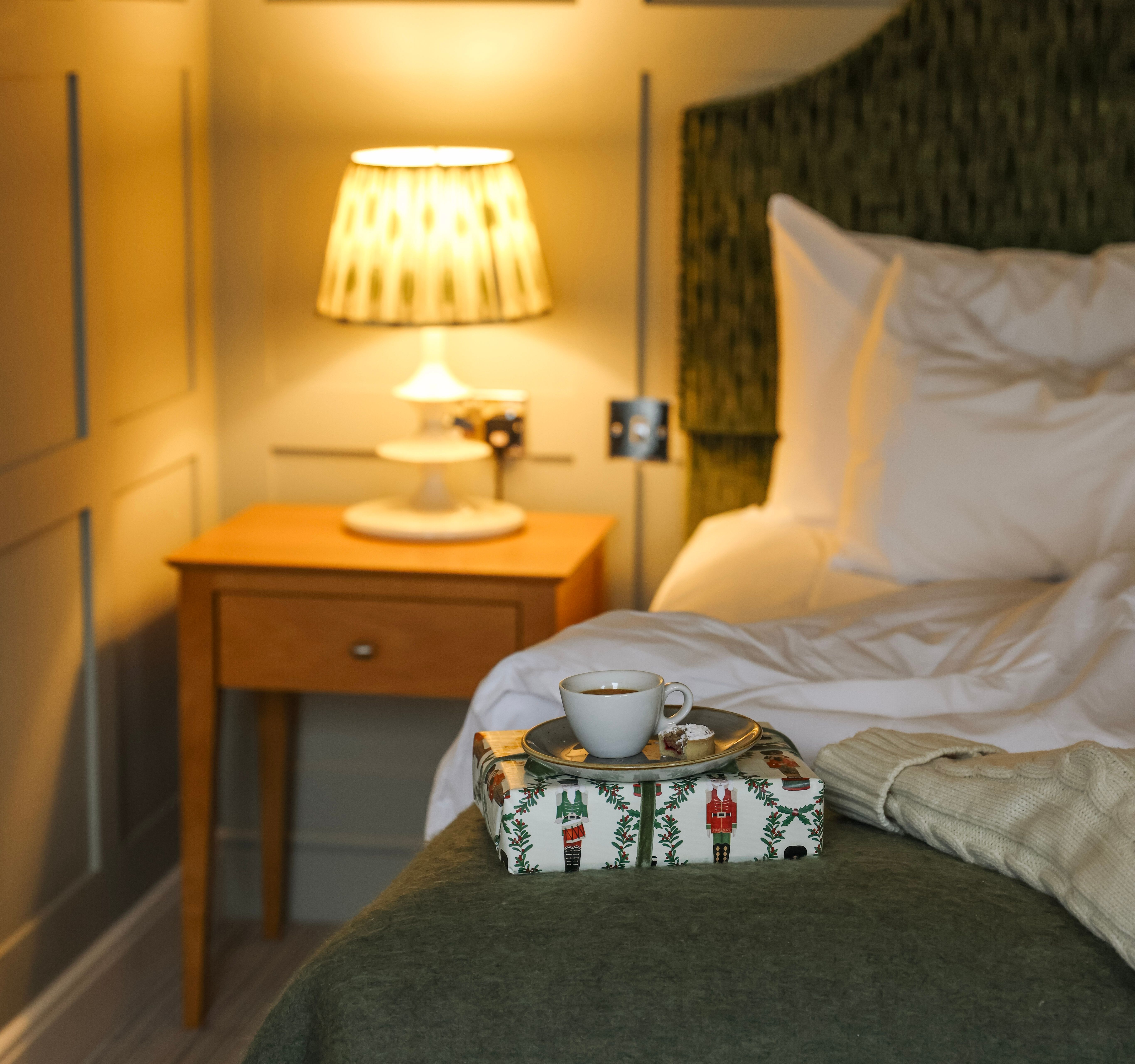 Cozy bedroom scene with a bedside lamp, a cup of coffee on a wrapped gift, and a bed with white pillows and a green throw.